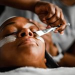 Image depicts Black woman getting a relaxing skincare treatment with esthetician.